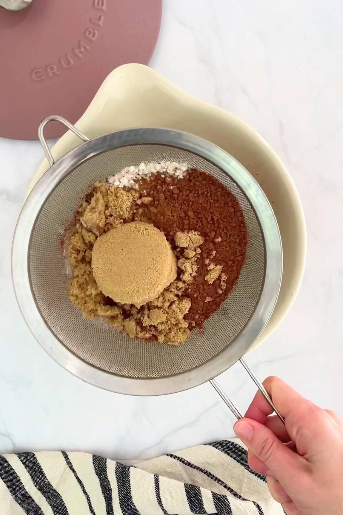 Hand holding a mesh sieve filled with brown sugar, cocoa powder and flour above a mixing bowl.