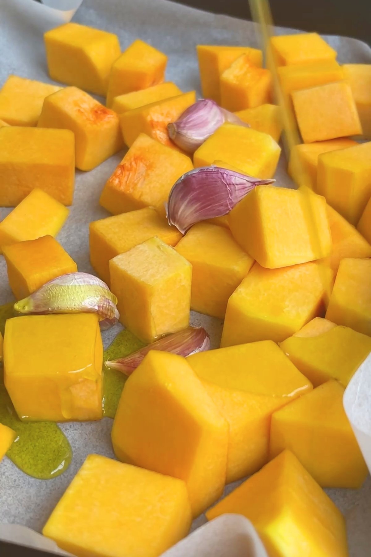 Raw pumpkin cubes and unpeeled garlic cloves on a parchment-lined tray, drizzled with olive oil.