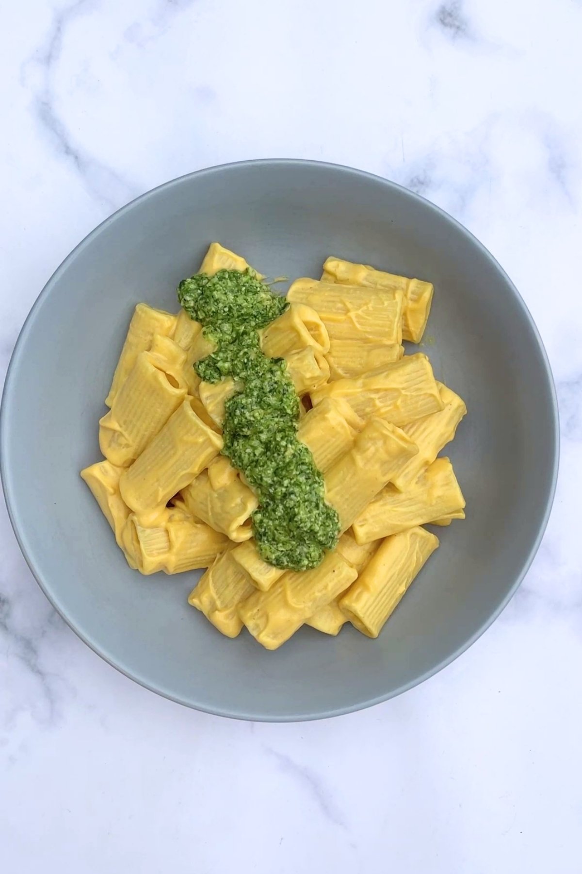 Creamy rigatoni pasta served in a grey bowl, topped with a generous spoonful of green basil pesto.