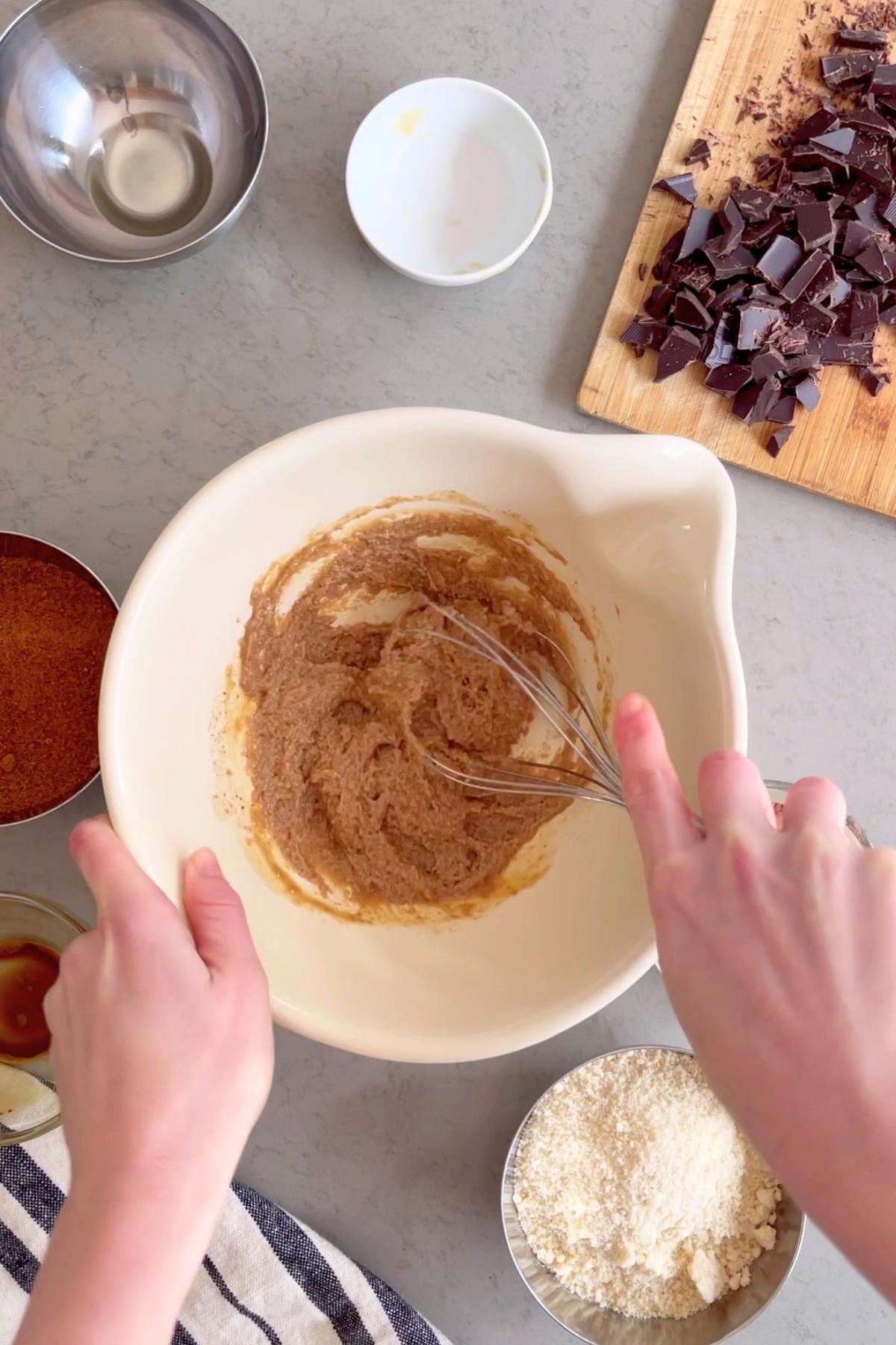 Hands whisk batter in white bowl, with chopped chocolate and small bowls nearby.