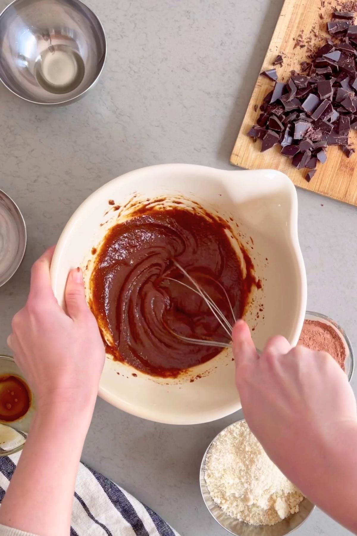 Hands whisk smooth chocolate batter in a white bowl, with chopped chocolate on a board and metal bowls nearby.