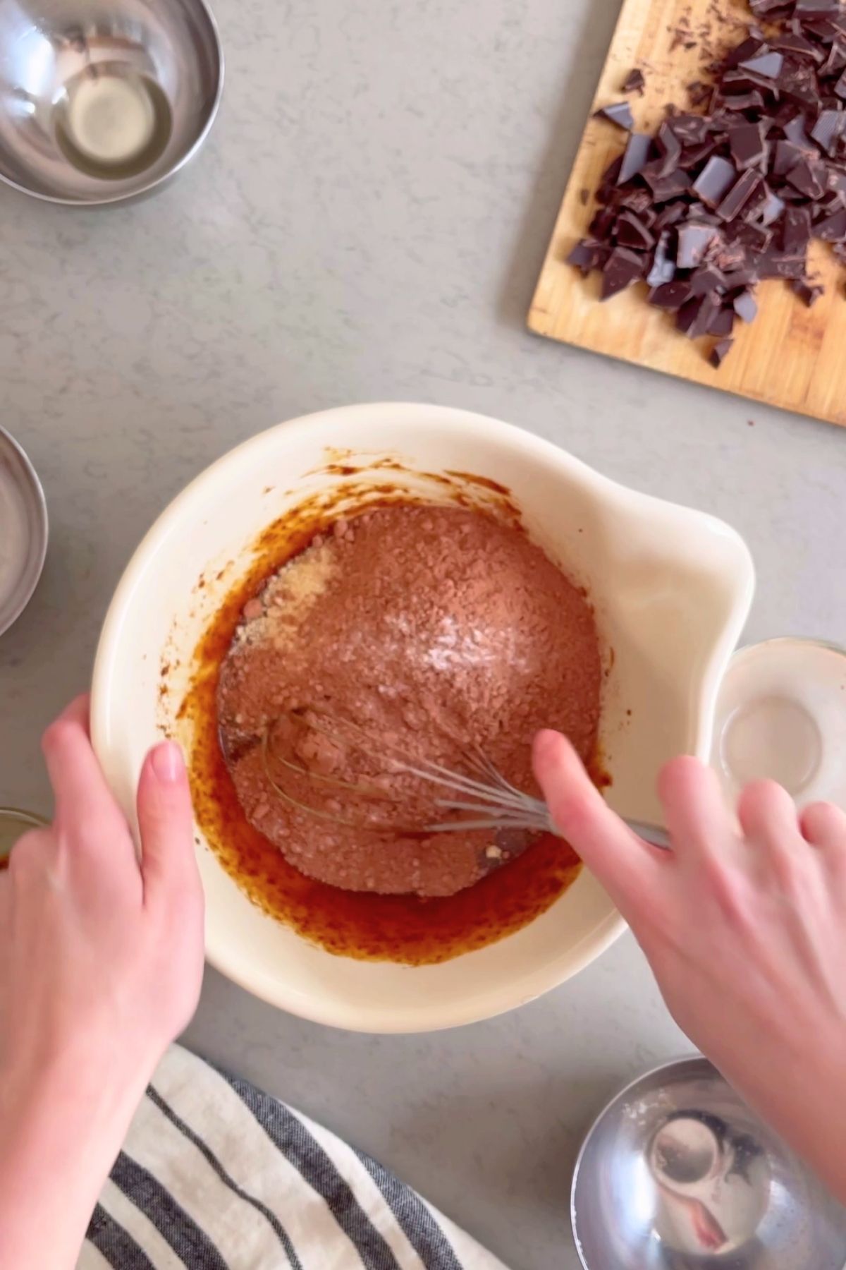 Hands whisk batter in a white bowl with chopped chocolate on a wooden board nearby.