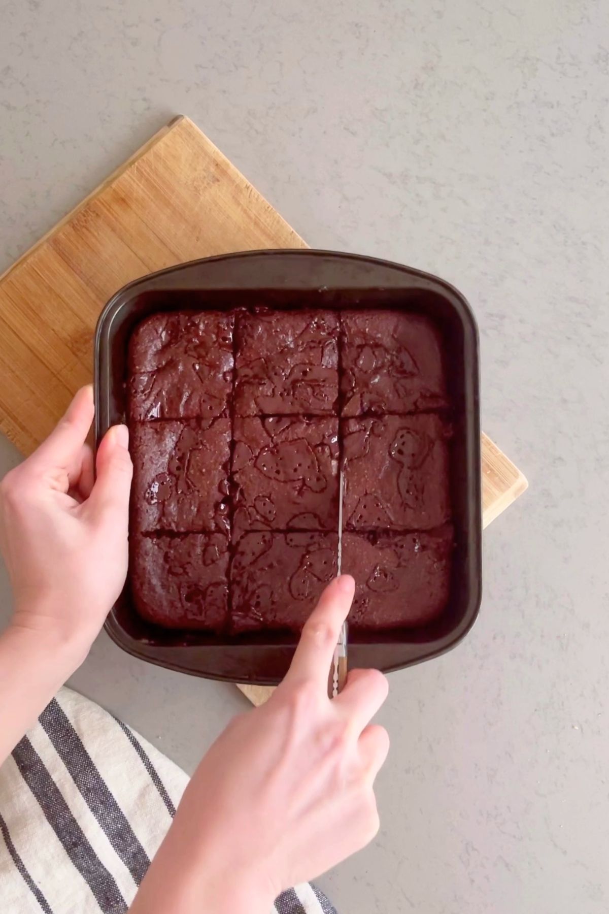 Hands slicing baked brownies into even squares inside a square pan.