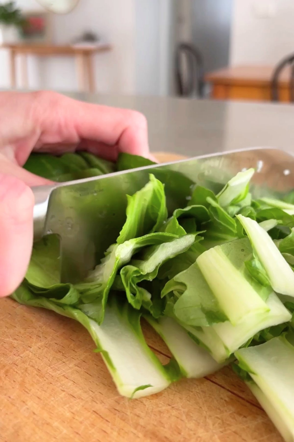 Hand slicing fresh bok choy leaves on a wooden chopping board with a sharp kitchen knife.