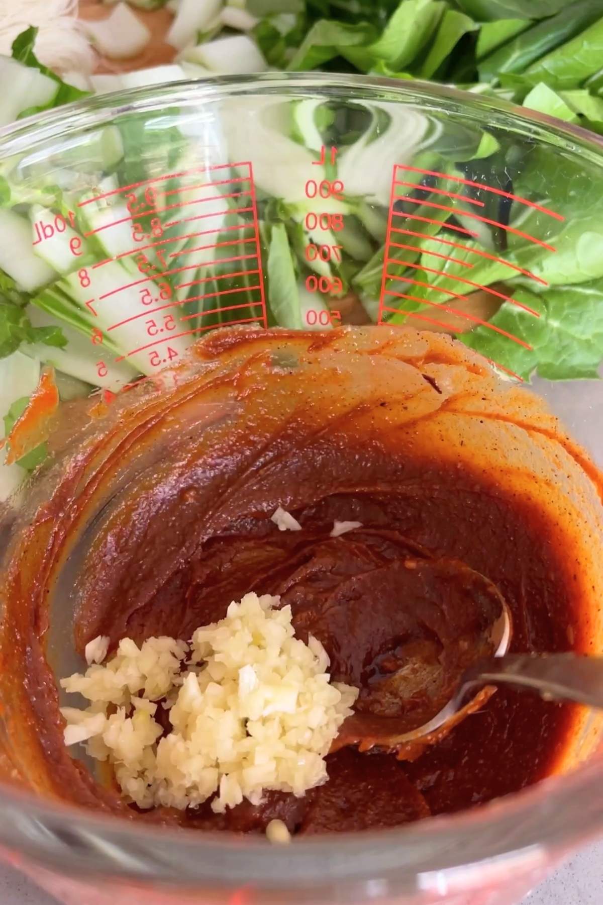 Minced garlic added to a thick reddish stock paste in a glass measuring jug, with chopped bok choy in the background.