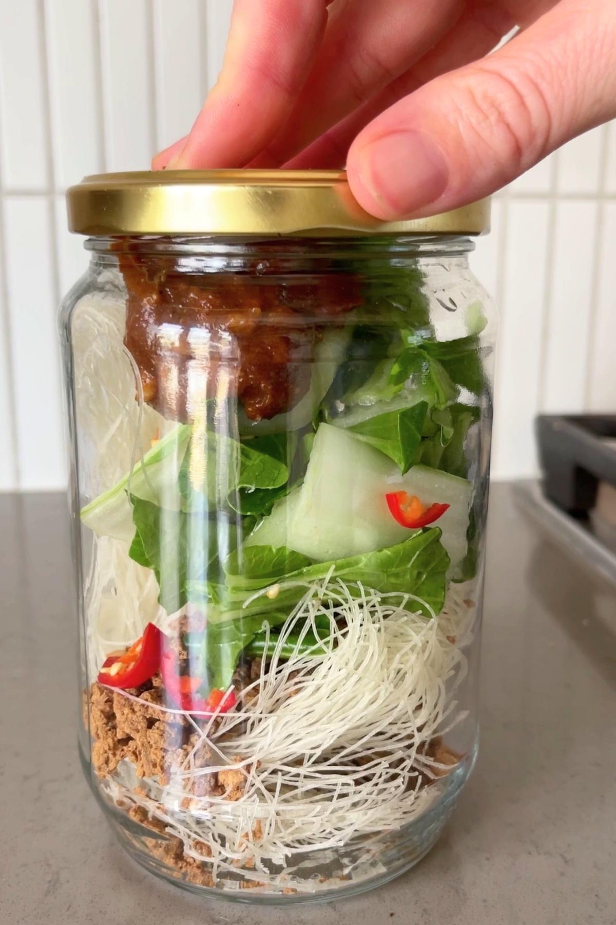 Layered jar of instant noodles with dry rice vermicelli, chopped bok choy, red chilli slices, TVP, and stock paste, being sealed with a gold lid.