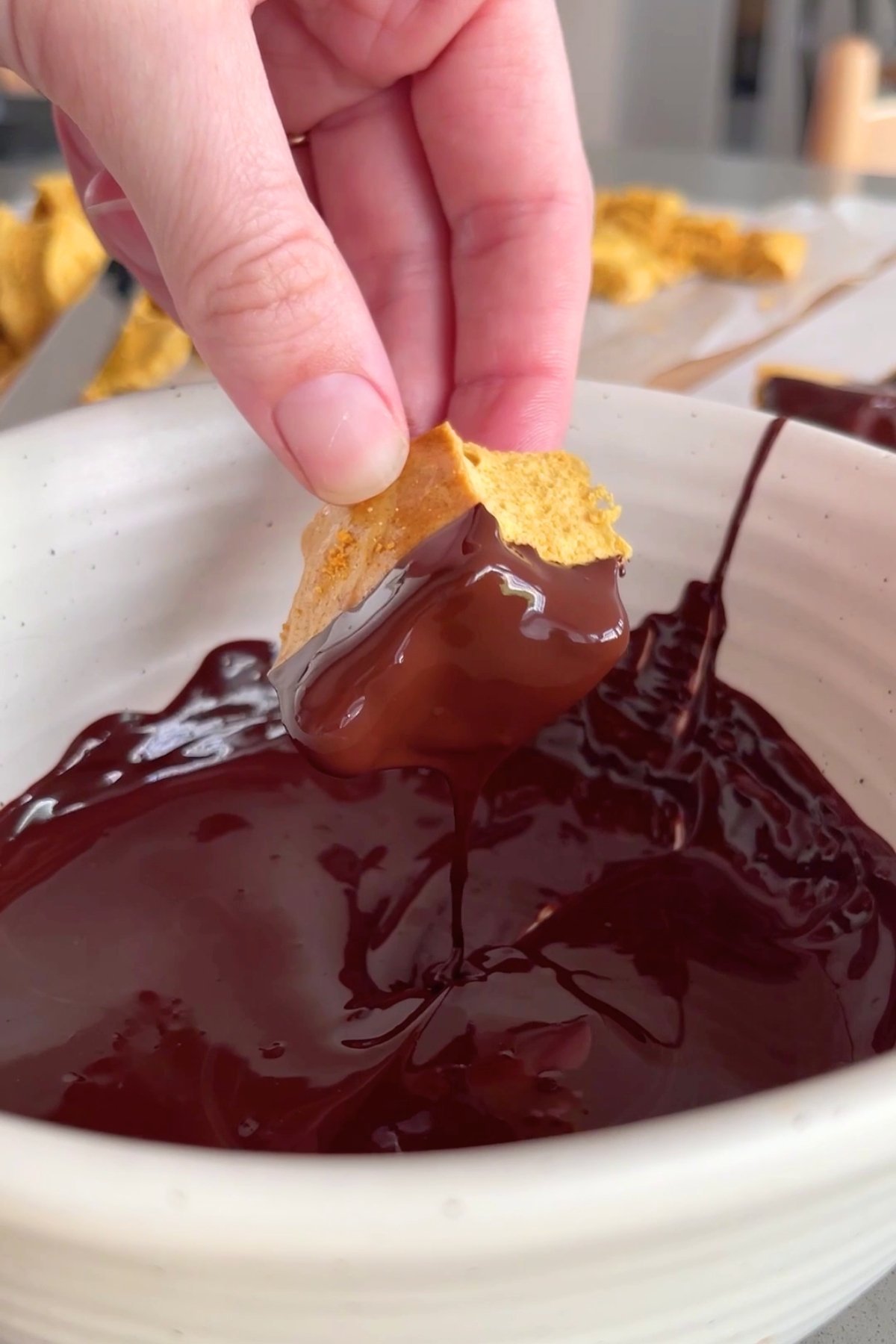 A hand dips a piece of golden honeycomb into a bowl of glossy melted dark chocolate.