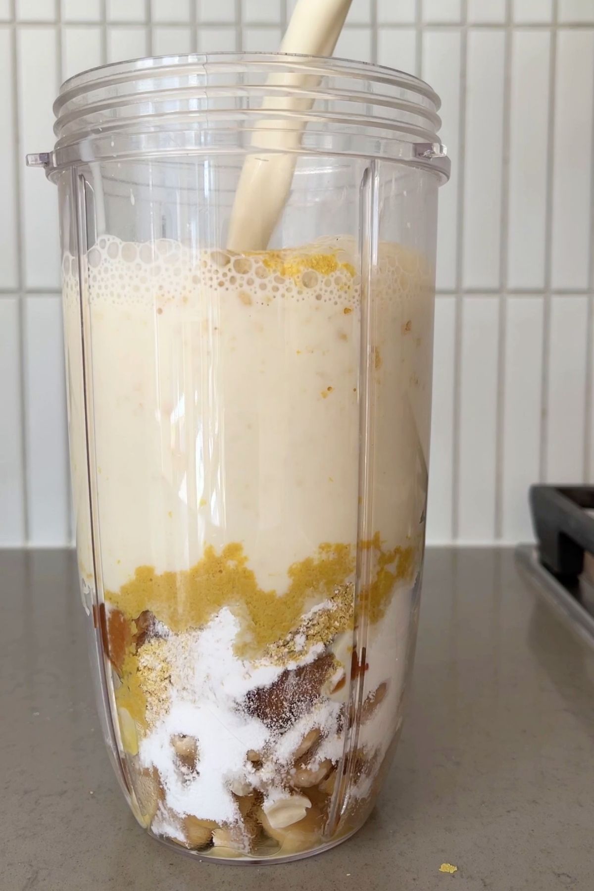 Unblended cashews, seasonings, and soy milk layered in a clear blender cup on a kitchen counter.