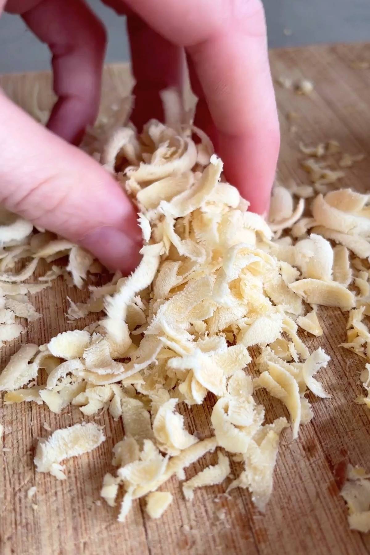Hand picking up soft, pale yellow shreds of vegan cheese from a wooden board.