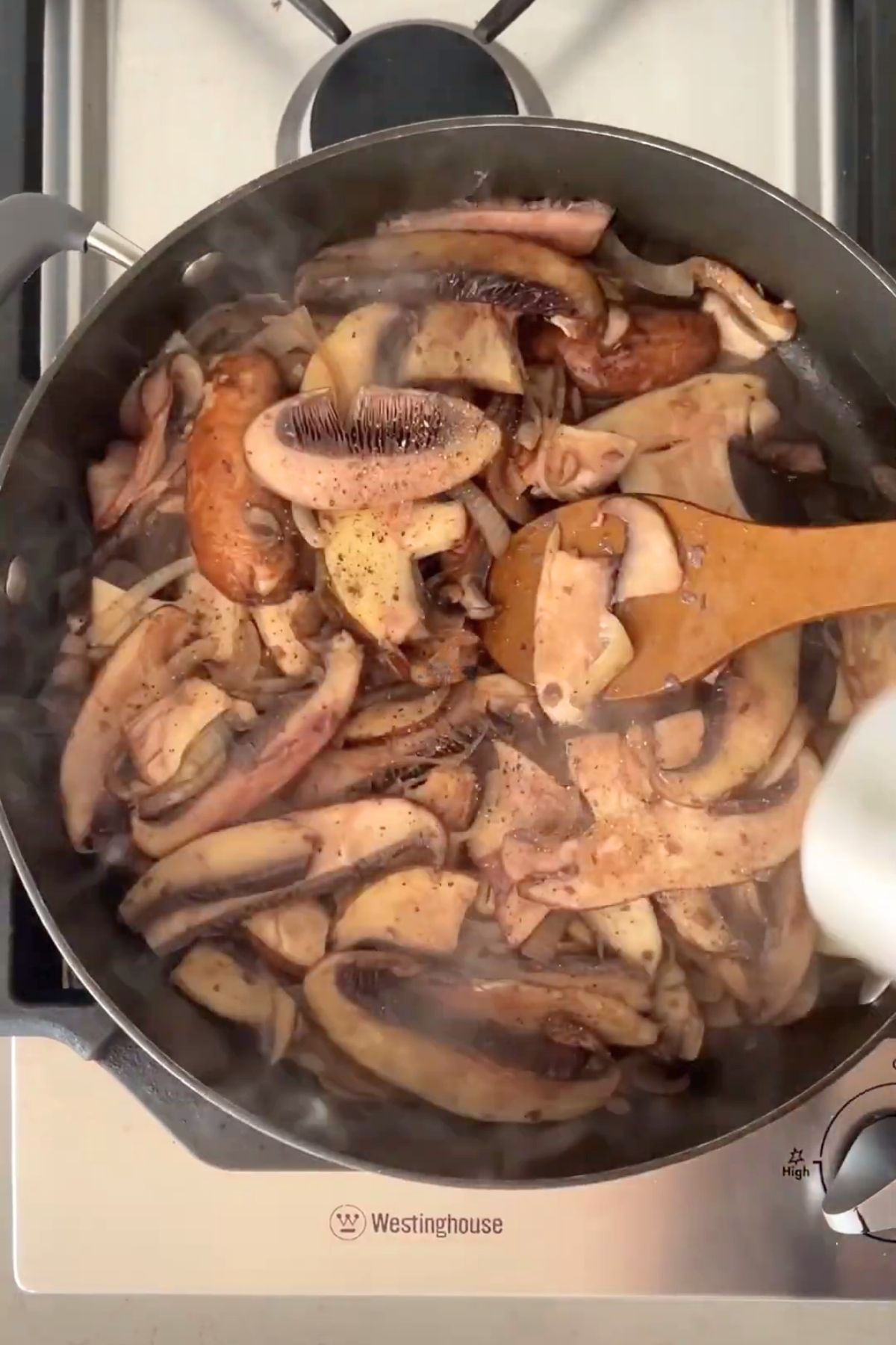 Sliced mushrooms and onions cooking in a pan, with steam rising and a wooden spoon stirring through the mixture.