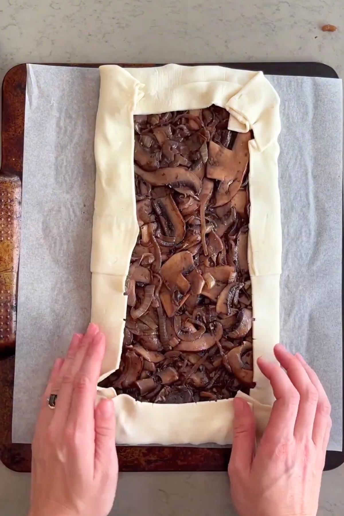 Hands folding the edges of puff pastry over a cooked mushroom and onion filling on a lined baking tray.