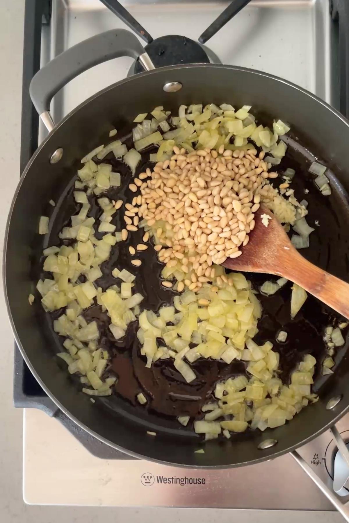 Diced onions sautéing in oil with a heap of pine nuts and minced garlic in a black pan on a stovetop.