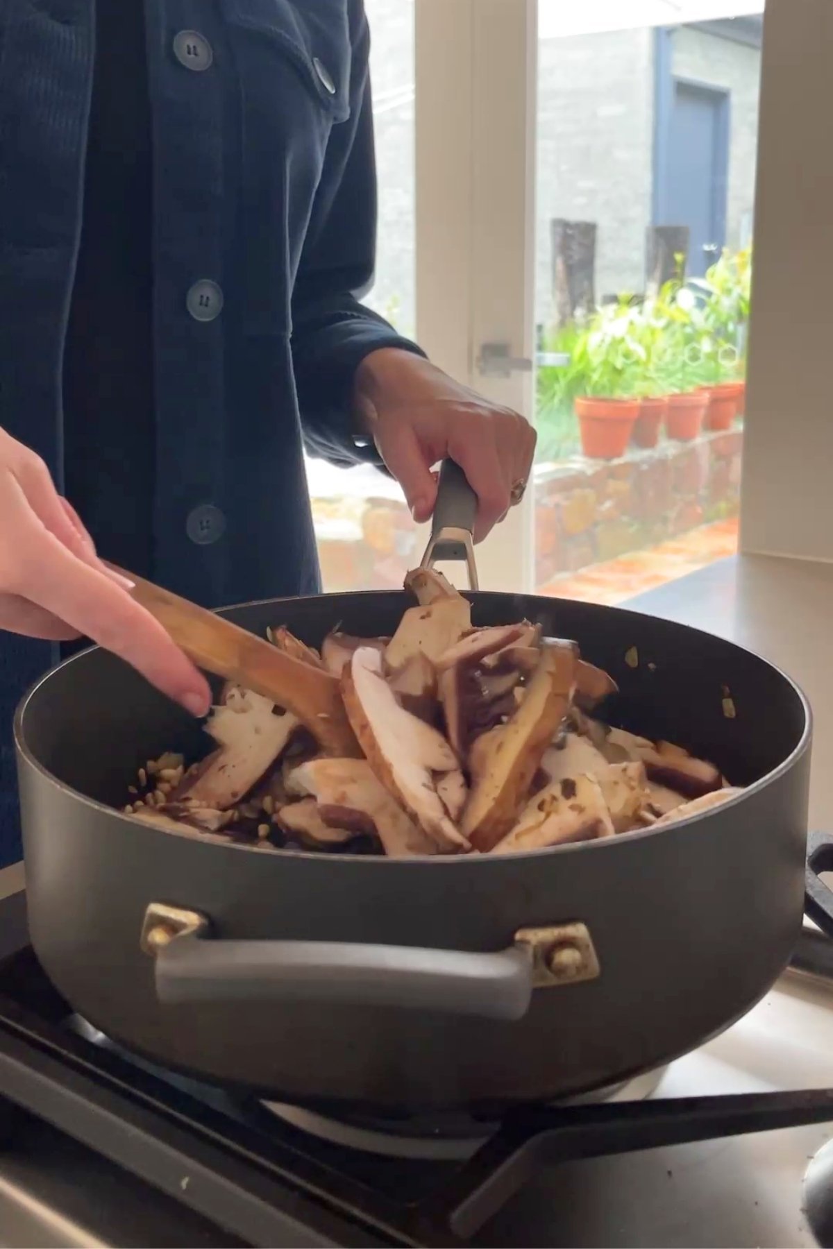 Sliced portobello mushrooms being stirred into a pan by hand over sautéed onions and pine nuts.