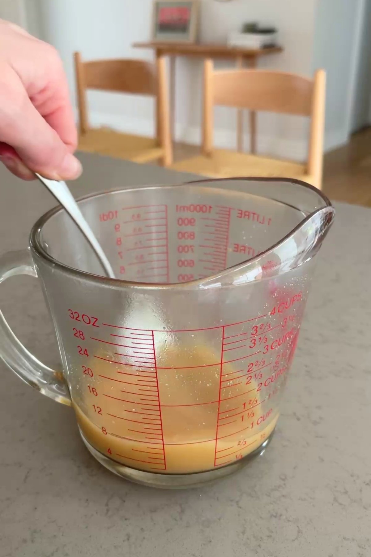 Brownish liquid being stirred with a spoon in a glass measuring jug, placed on a kitchen counter.