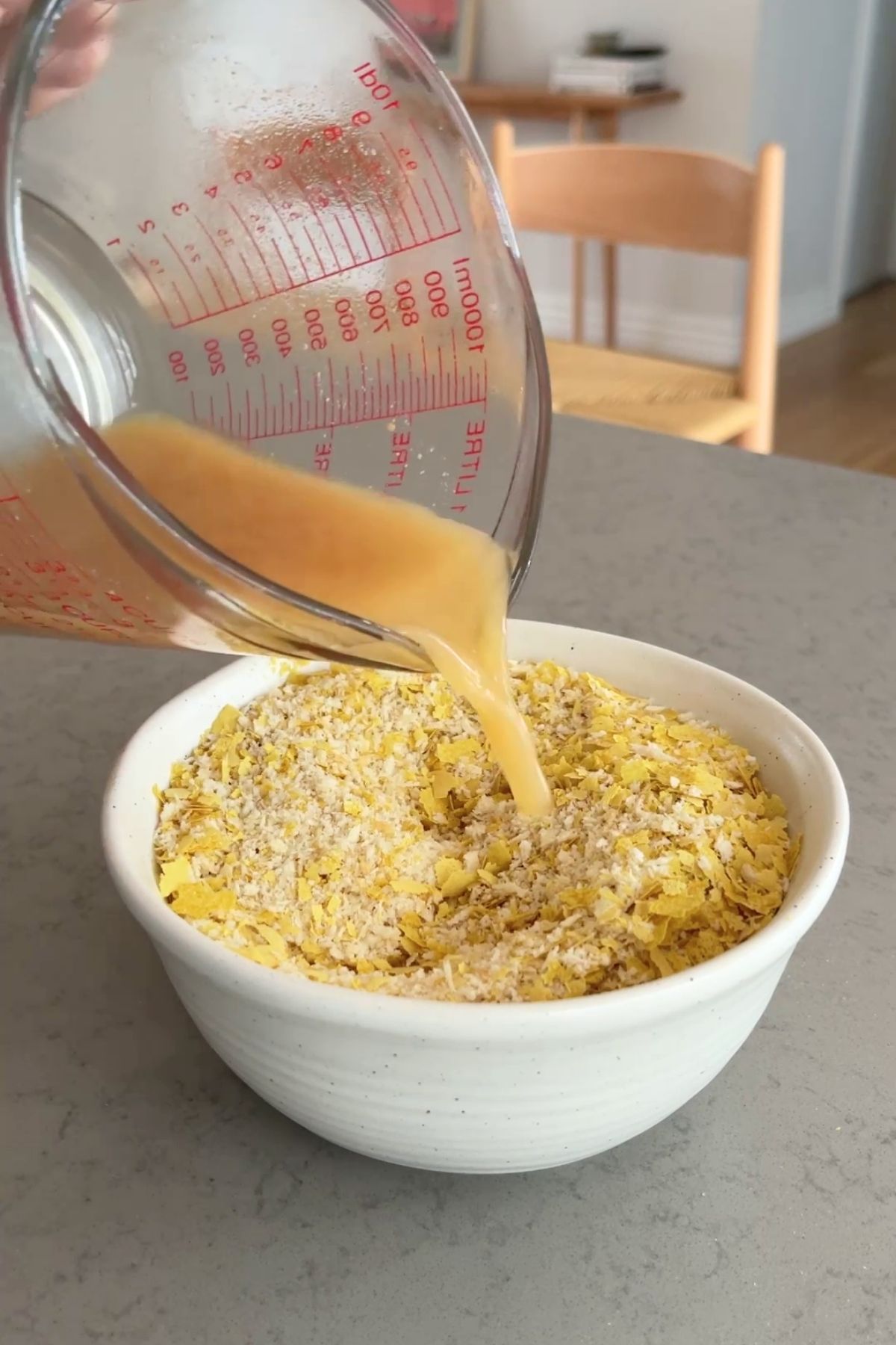 Golden liquid being poured into a white bowl filled with yellow nutritional yeast flakes and pale breadcrumbs.
