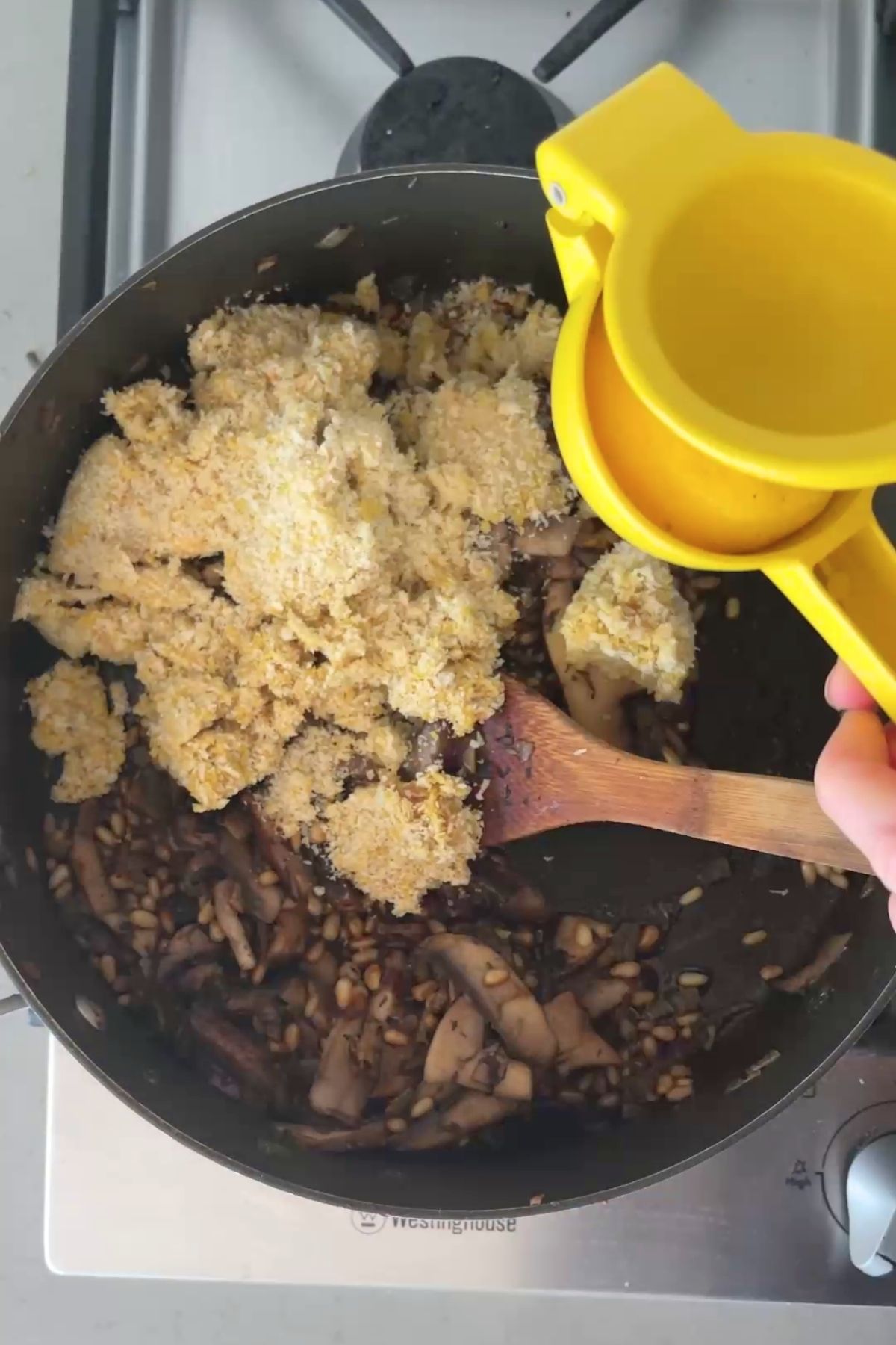 Soaked breadcrumb and nutritional yeast mixture being added to sautéed mushrooms and pine nuts, with a yellow citrus press held above.