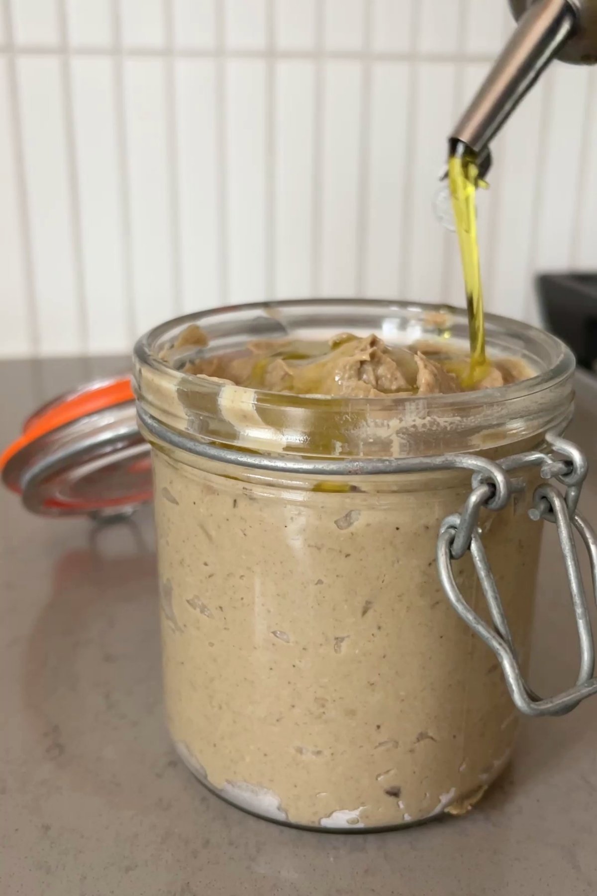 Olive oil being poured over creamy mushroom pâté in a glass jar with a metal clasp and orange seal.