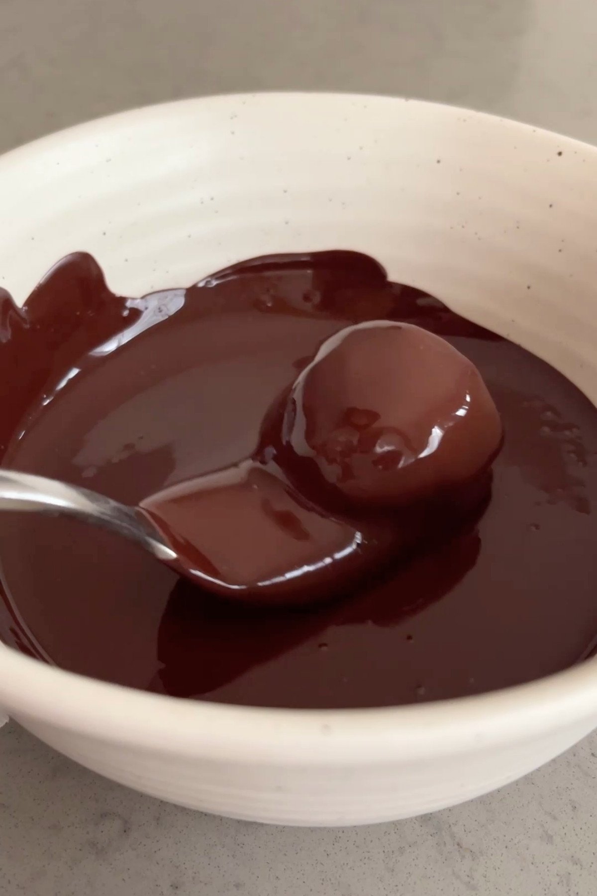 A truffle ball being coated in glossy melted dark chocolate with a spoon in a white bowl.