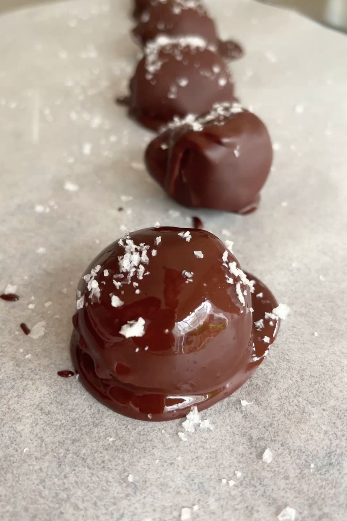 Glossy dark chocolate truffles topped with flaky sea salt, set in a neat row on parchment paper.