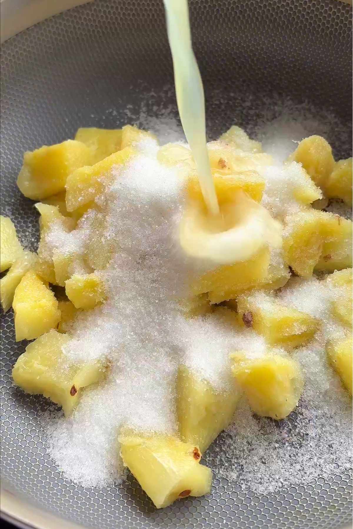 Pineapple chunks and sugar in a pan as a pale liquid streams over them.