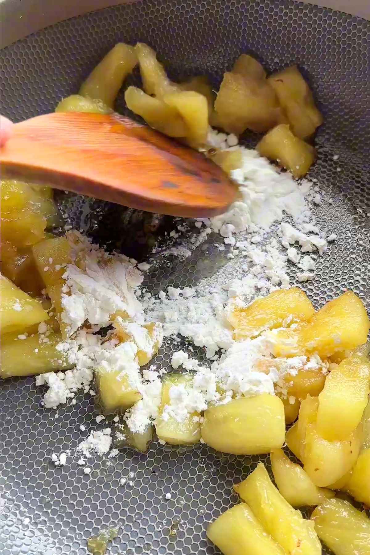 Cooked pineapple being stirred with cornstarch in a pan using a wooden spatula.