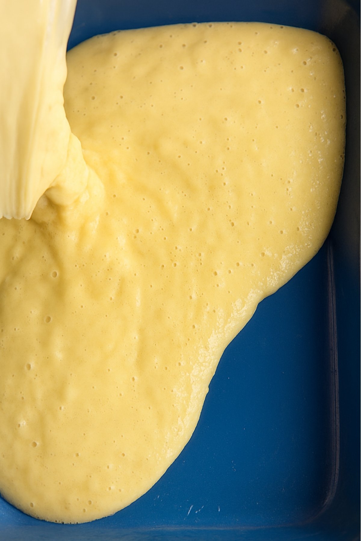 Creamy yellow mixture pours into a blue pan, bubbles forming.