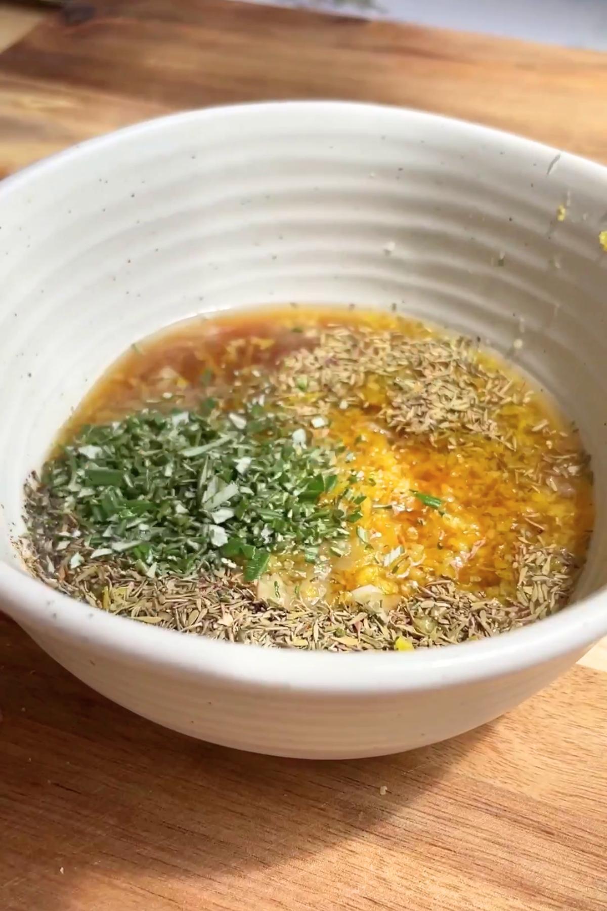 A white bowl filled with a lemon-colored liquid, topped with minced herbs and visible flecks of dried rosemary and thyme.
