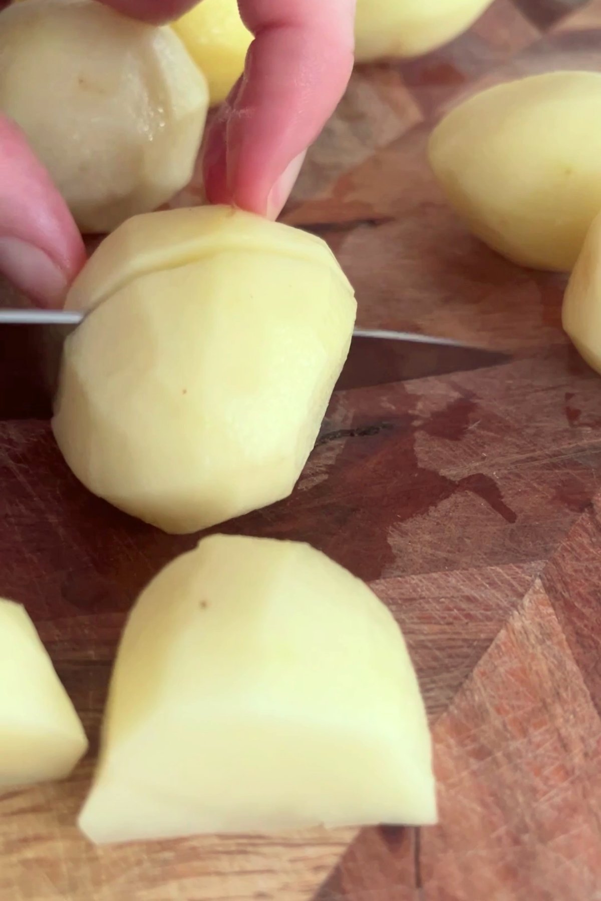 Peeled potato being sliced on a wooden chopping board with other cut pieces nearby.