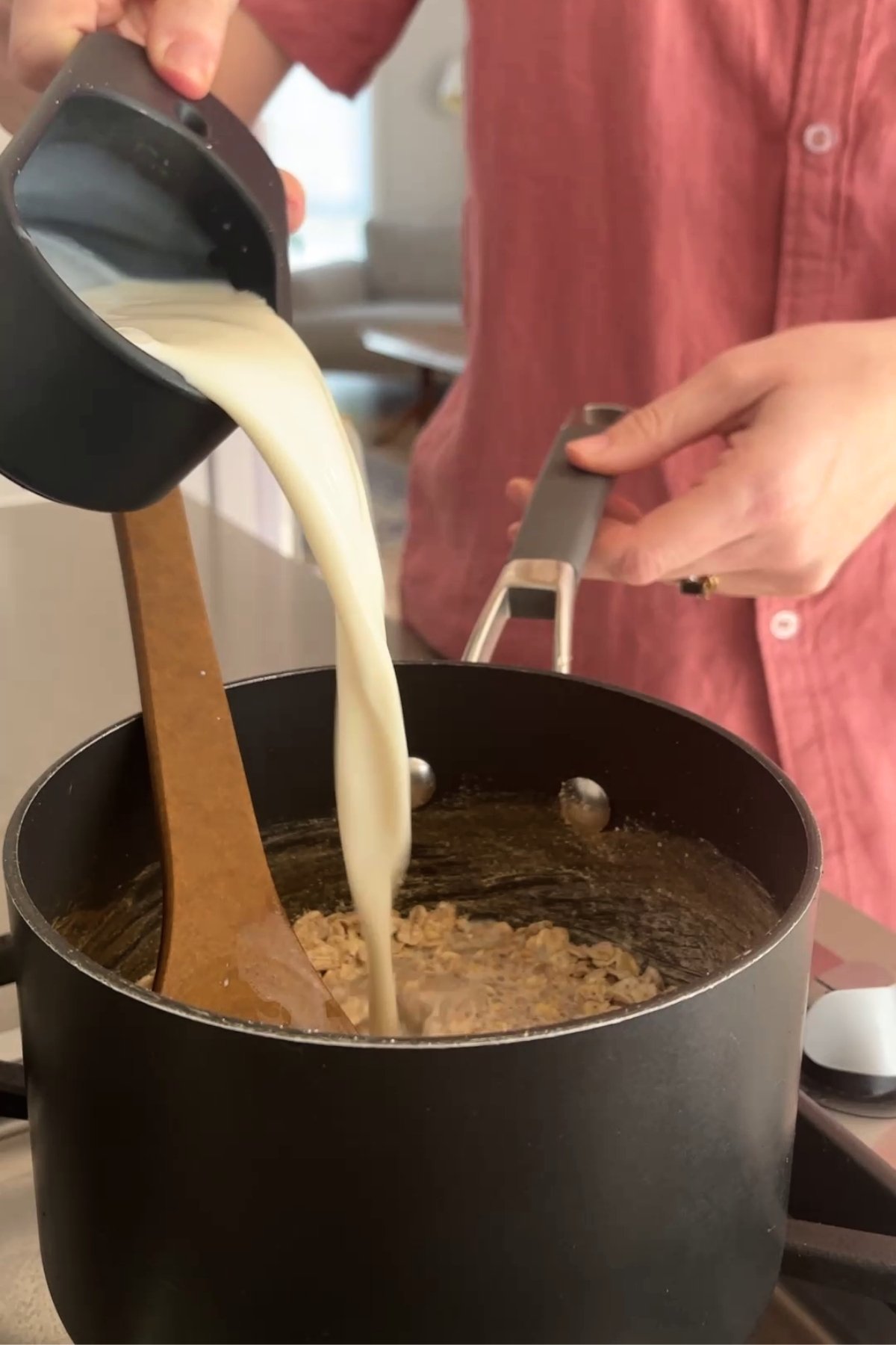 Soy milk being poured into a saucepan of oats and spices, with a wooden spoon resting inside.