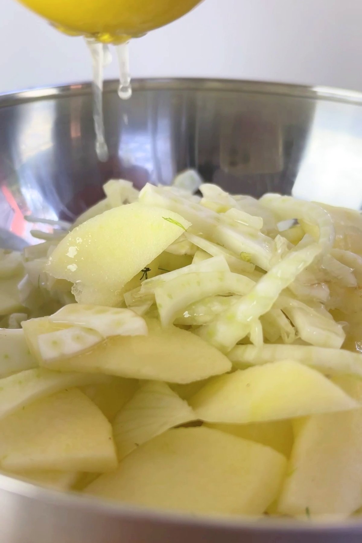 Lemon juice dripping over thinly sliced fennel and apple in a metal mixing bowl.