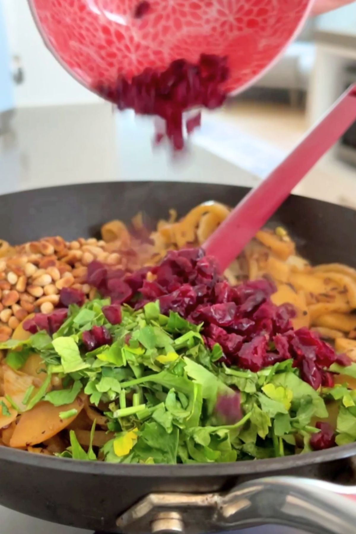 Chopped dried cranberries, fresh parsley, and toasted pine nuts being added to a pan with cooked vegetables.