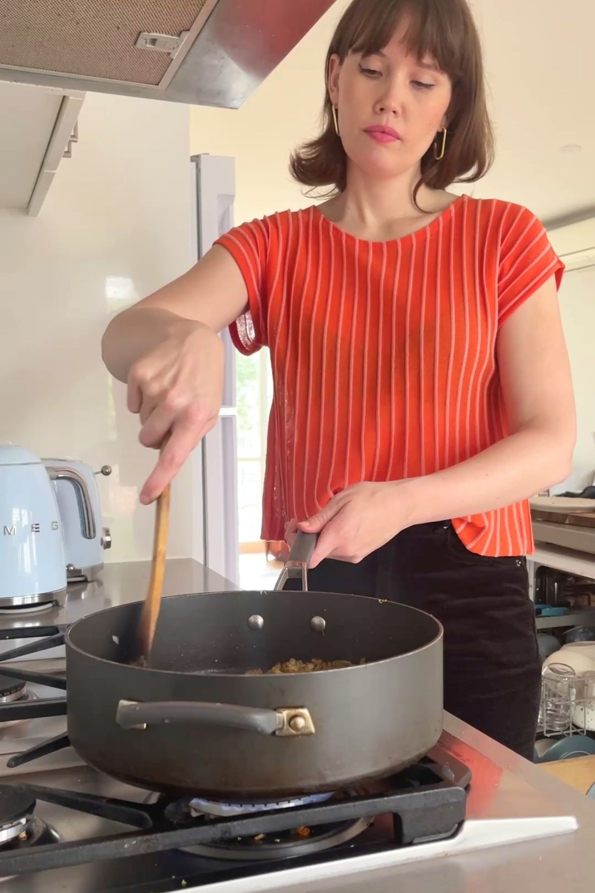 Woman (Liz Douglas) in an orange striped shirt stirring onions in a black saucepan over a gas flame on a stovetop.