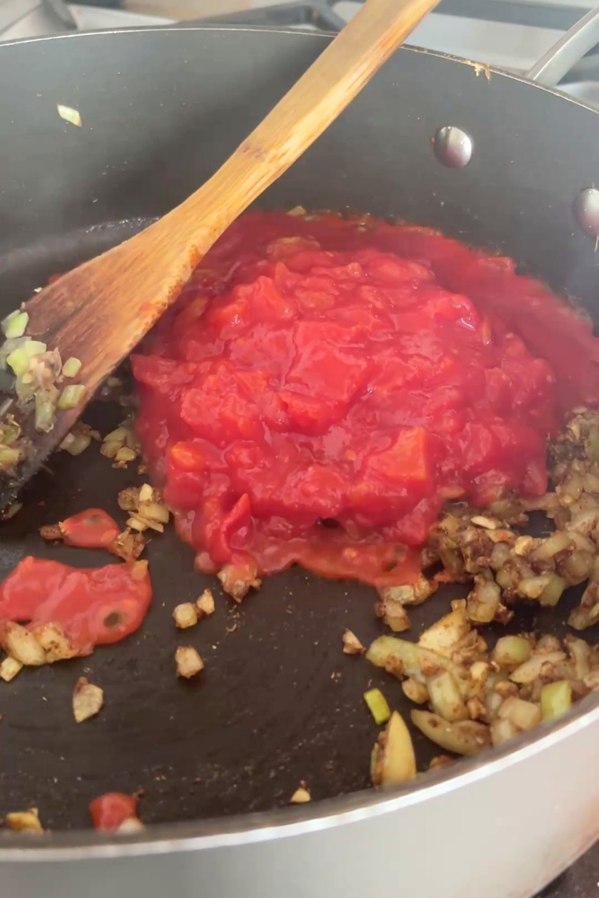 Chopped tomatoes added to sautéed onion and spices in a black saucepan, with a wooden spoon resting on the side.