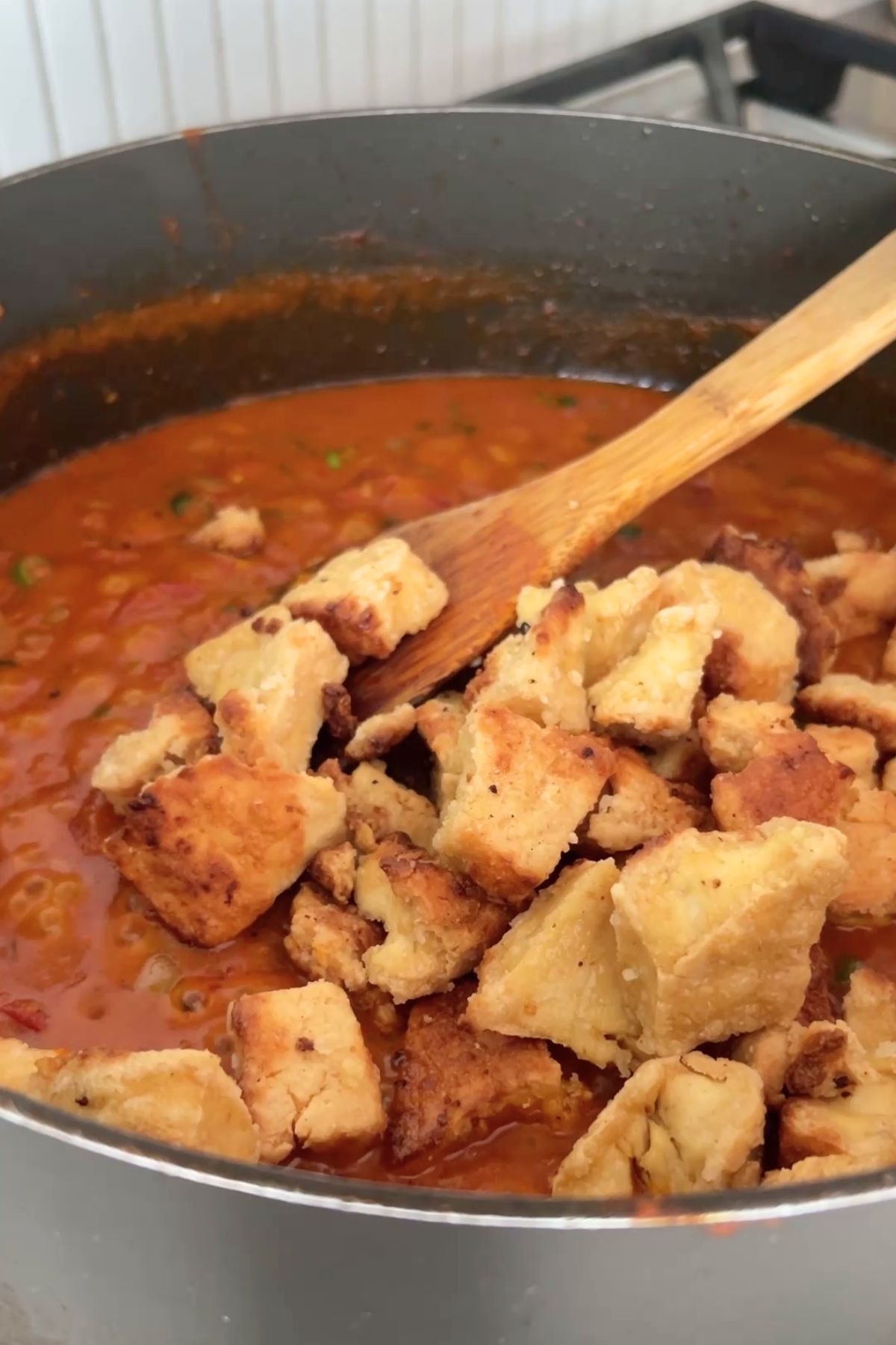 Golden, crispy tofu pieces being stirred into a rich, orange-red curry sauce with a wooden spoon in a black saucepan.