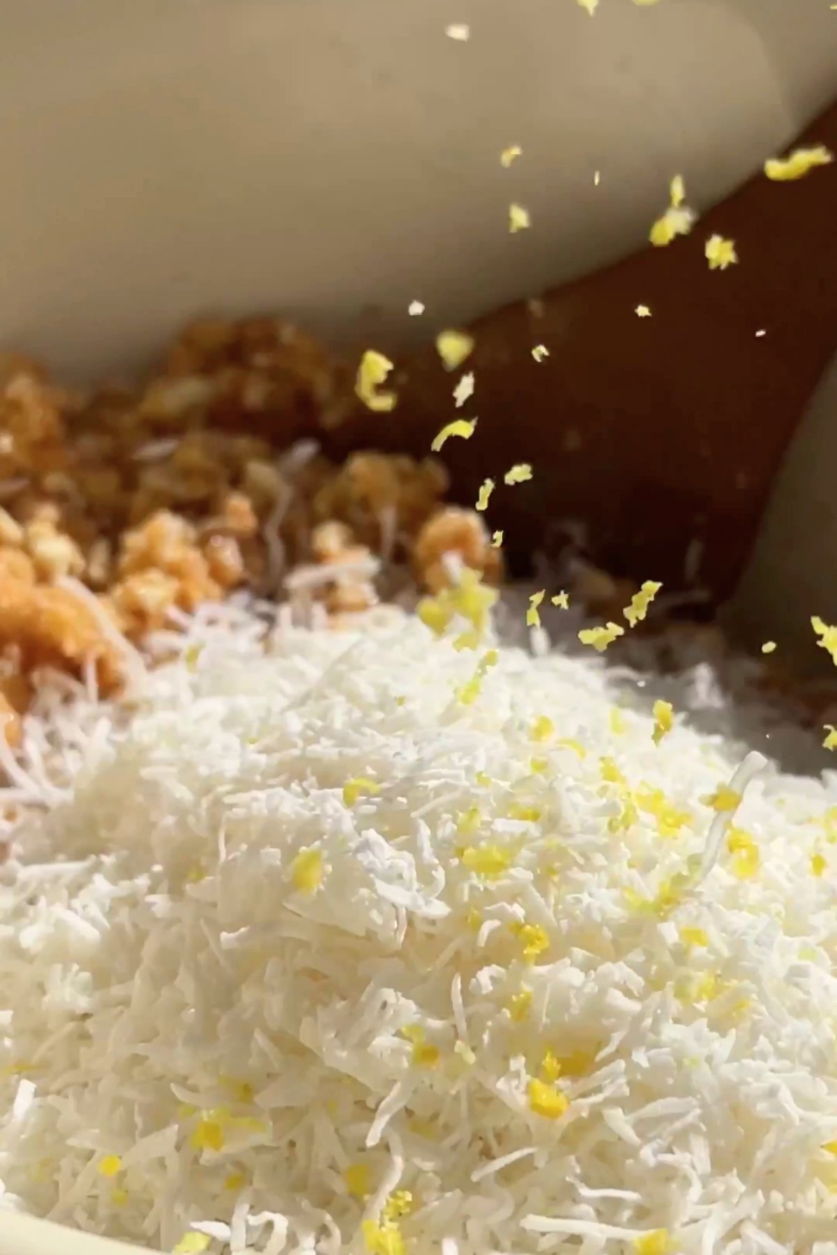 Lemon zest falls over shredded coconut and biscuit crumbs in a mixing bowl, ready to be combined.