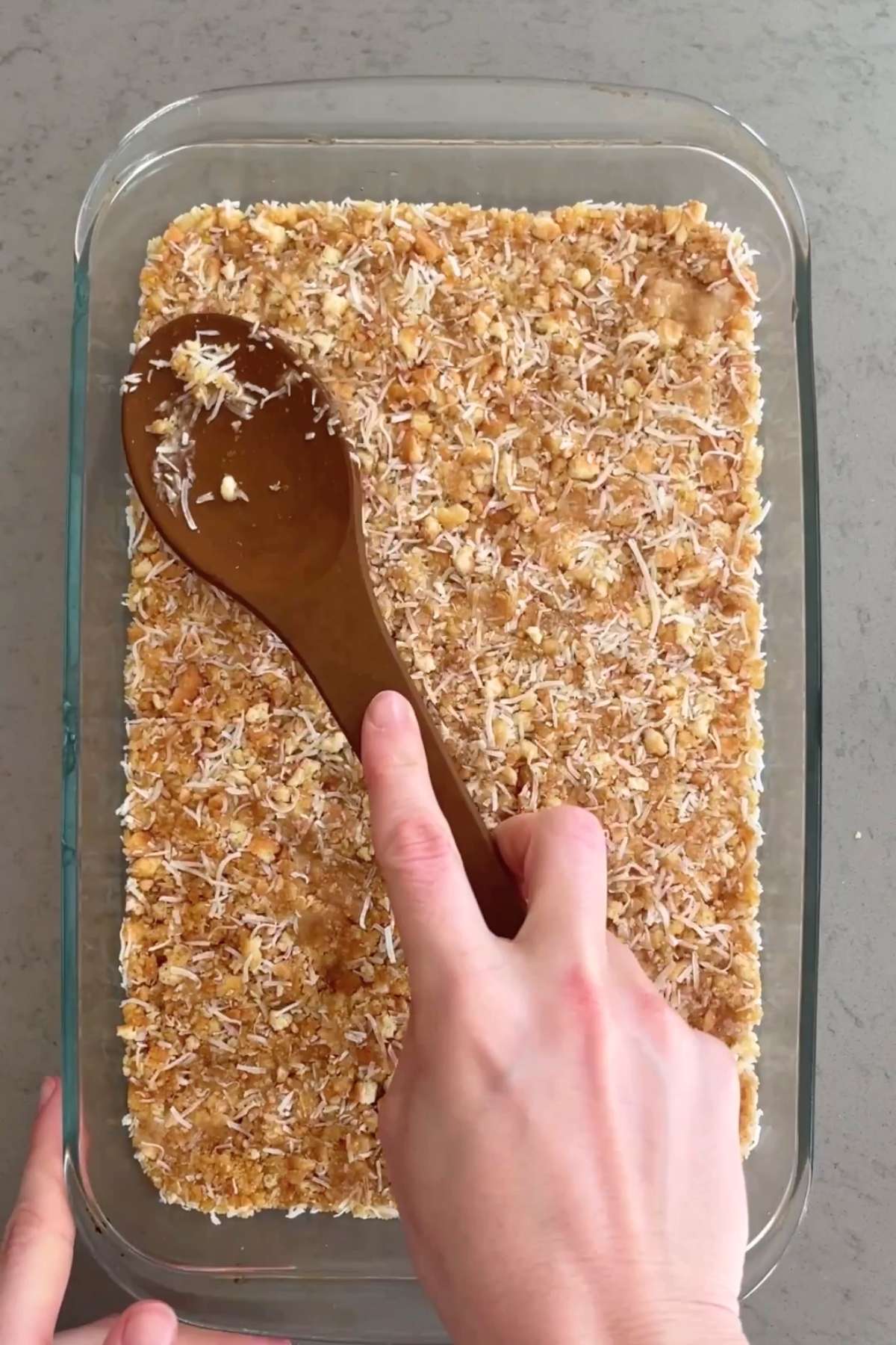 A wooden spoon presses the crumbly lemon slice mixture firmly into a glass tray to form the base layer.