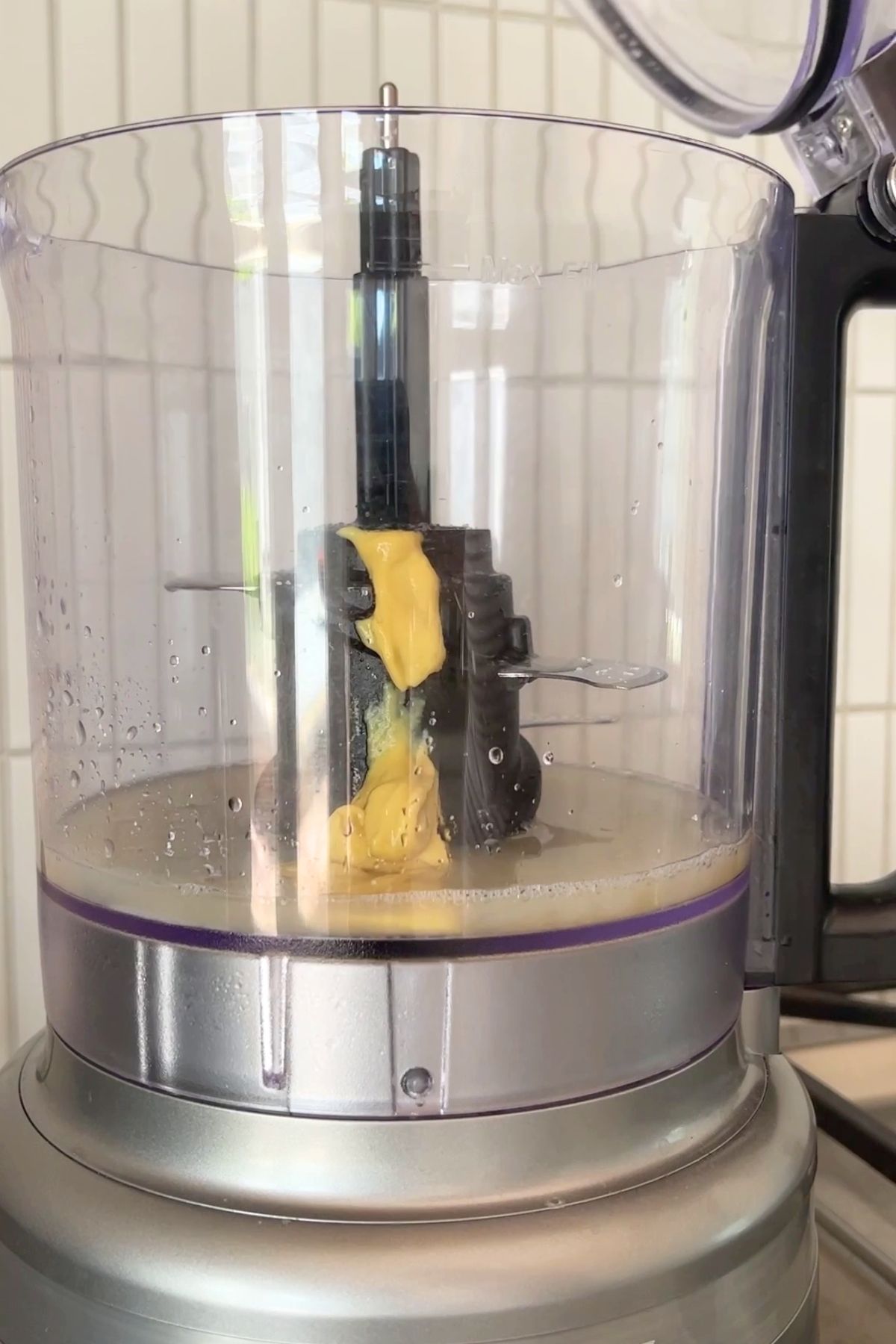 Thick mustard-colored mixture clings to the center blade of a food processor surrounded by aquafaba.