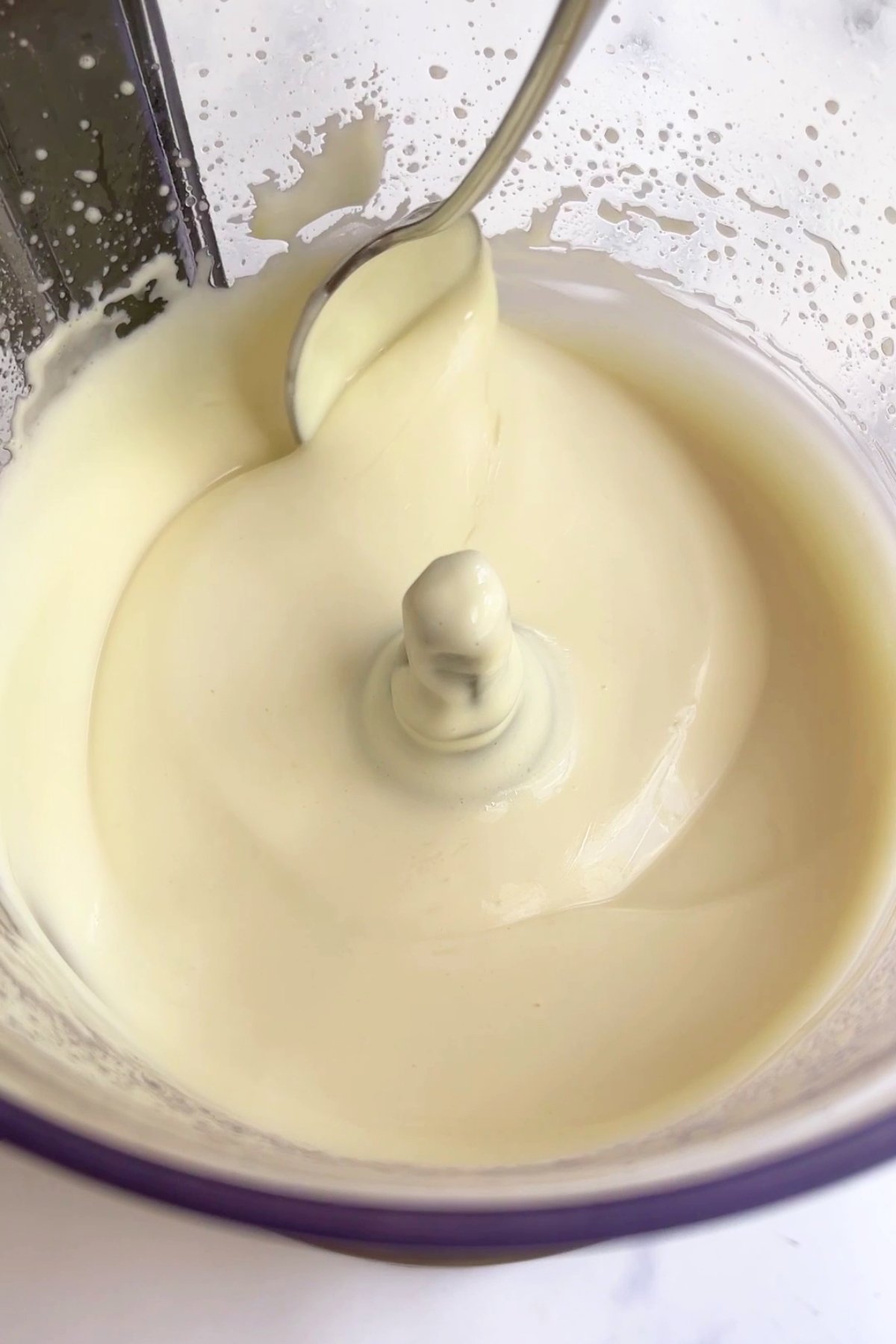 A spoon glides through thick, creamy white mixture inside a food processor bowl.