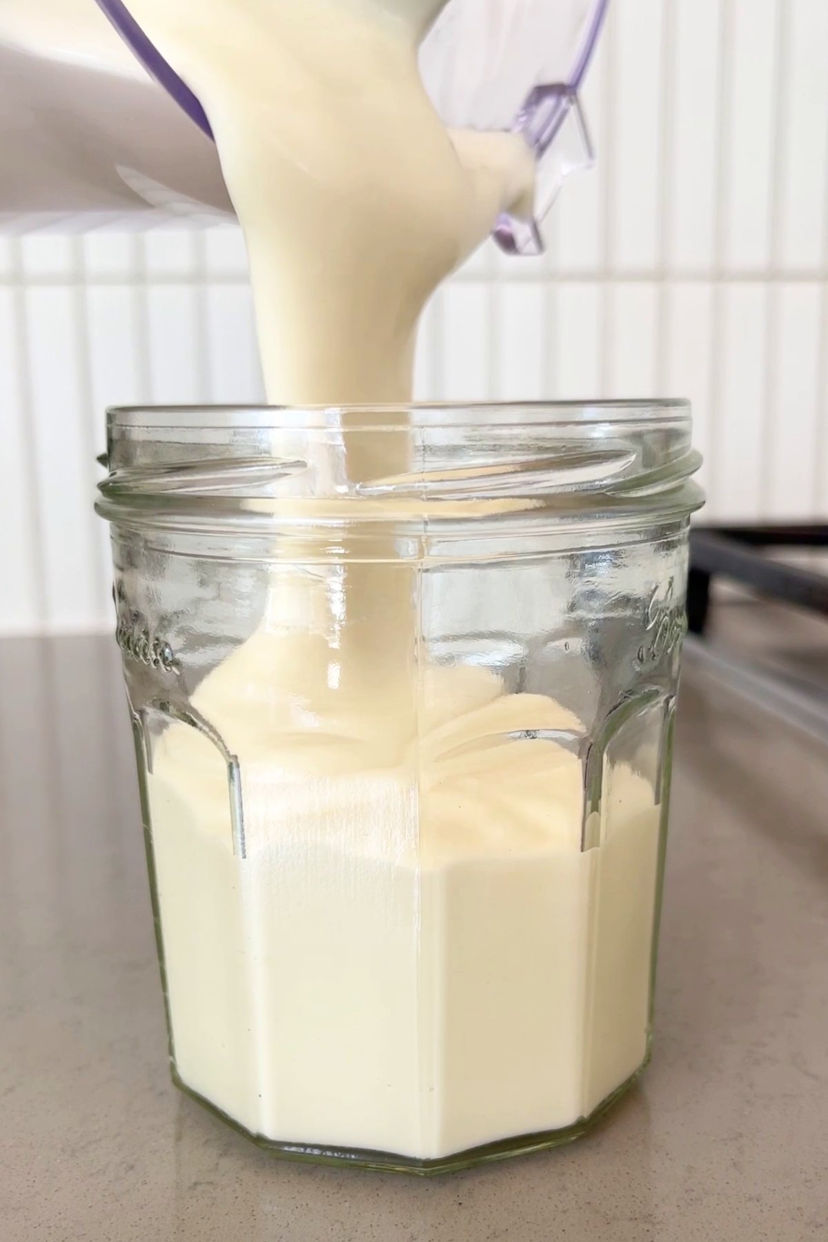 Thick, creamy mixture is being poured from a blender into a clear glass jar on a countertop.