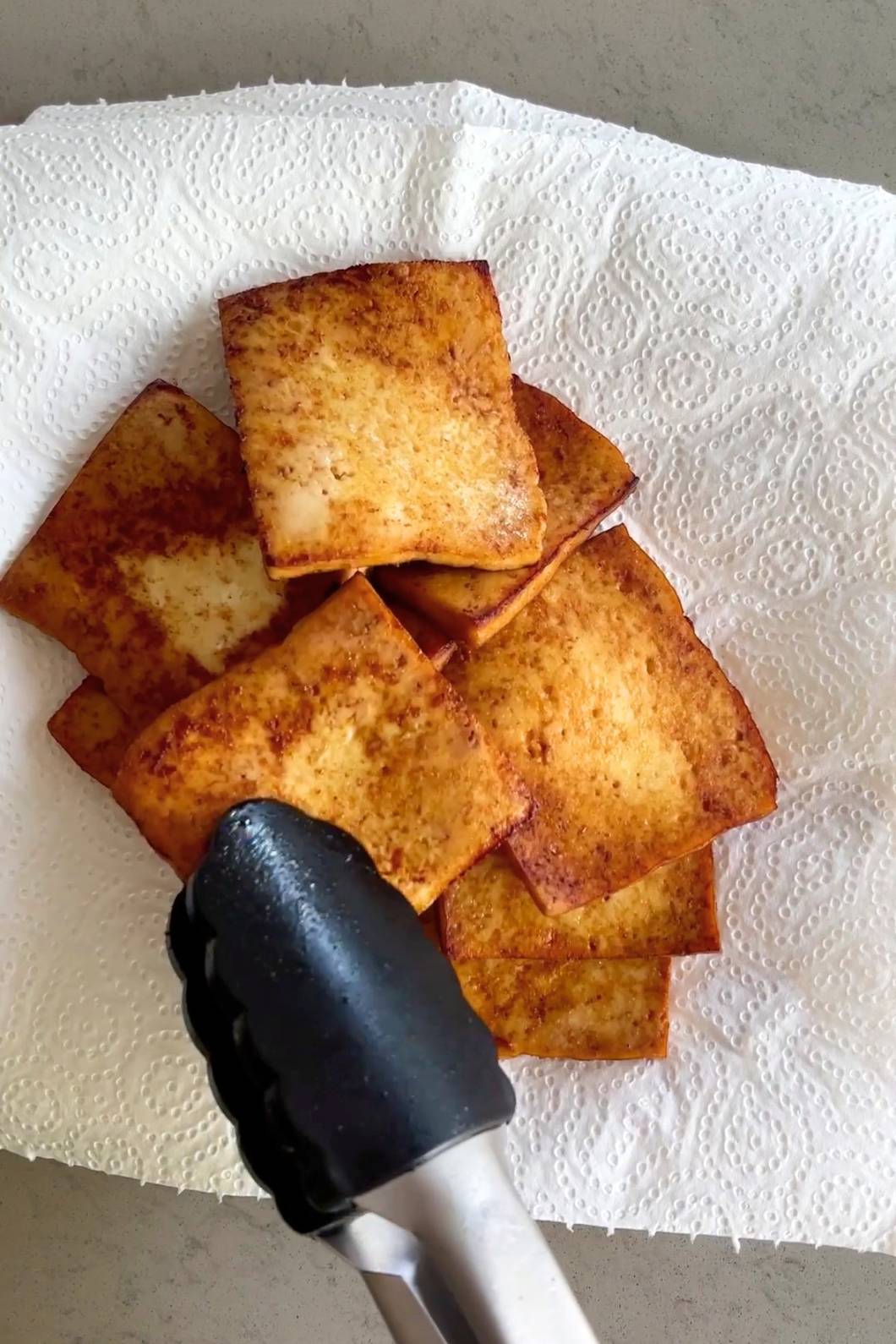 Square golden-brown tofu pieces draining on paper towel held by tongs.