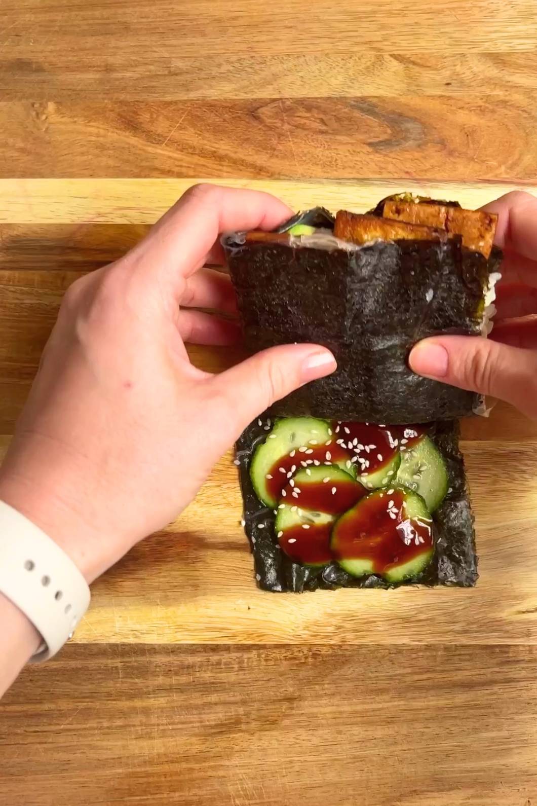 Hands folding a nori wrap over fillings on a wooden board.