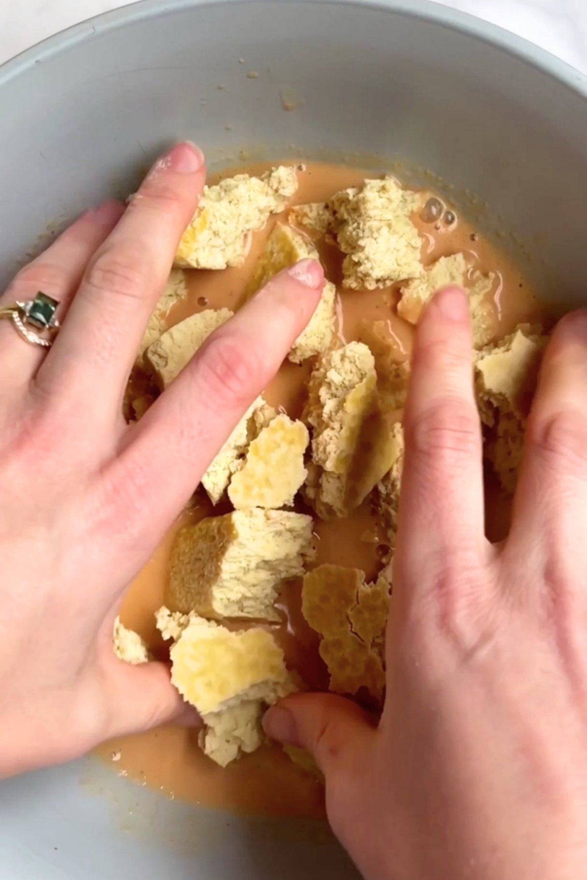 Hands mixing torn tofu chunks into a seasoned vegan buttermilk marinade in a blue bowl.