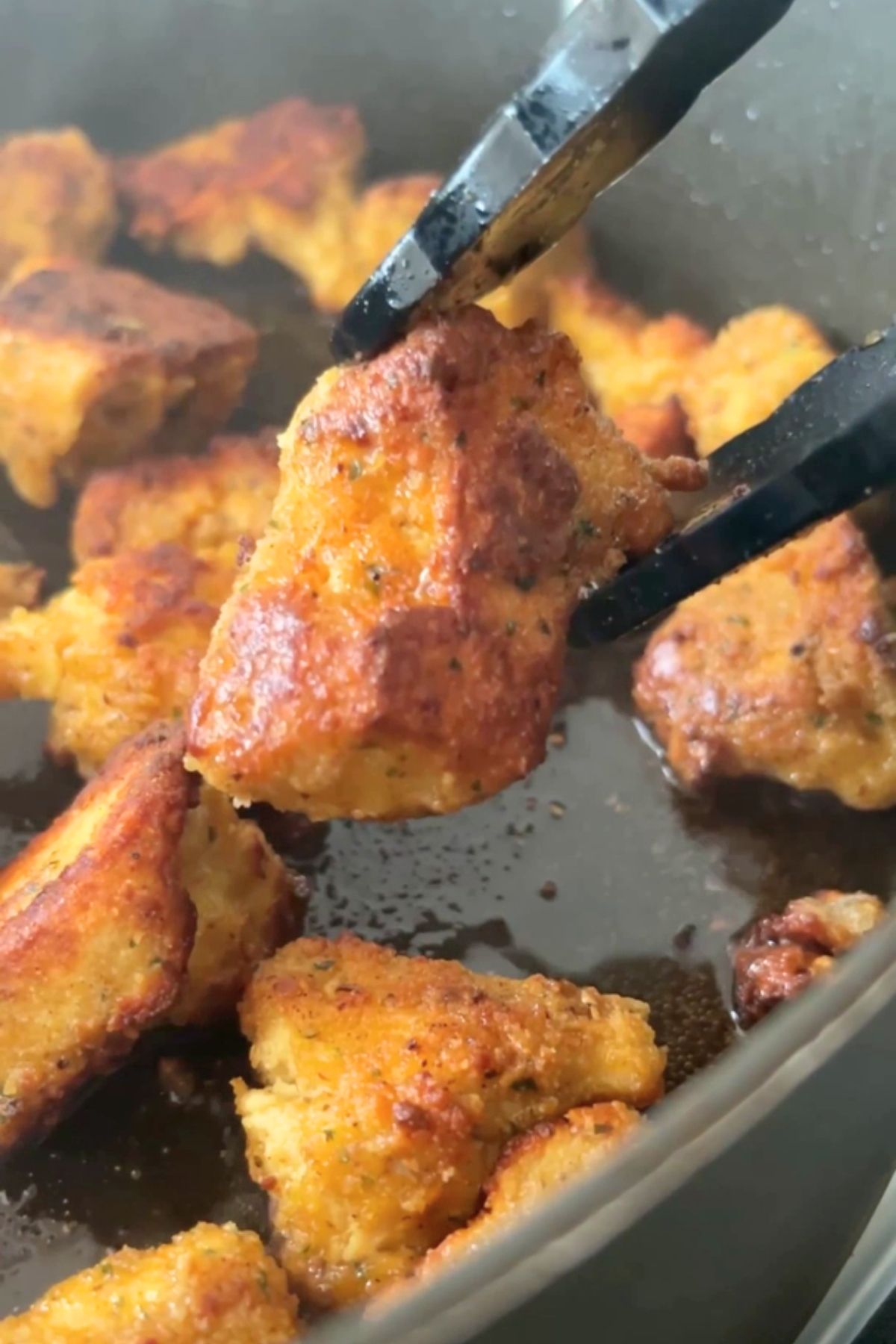 Crispy golden tofu piece being lifted from a hot pan with tongs, surrounded by sizzling fried chunks.