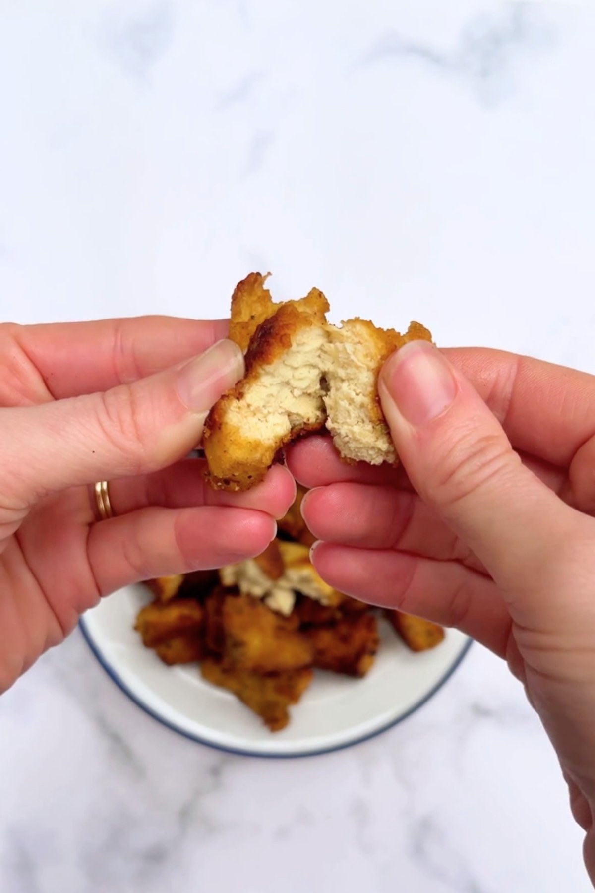 Two hands tearing open a crispy tofu piece, revealing its tender, layered, chicken-like texture inside.