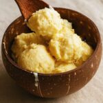 Wooden spoon lifts yellow ice cream from a coconut bowl as a drip runs down the rim.