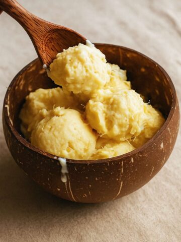 Wooden spoon lifts yellow ice cream from a coconut bowl as a drip runs down the rim.