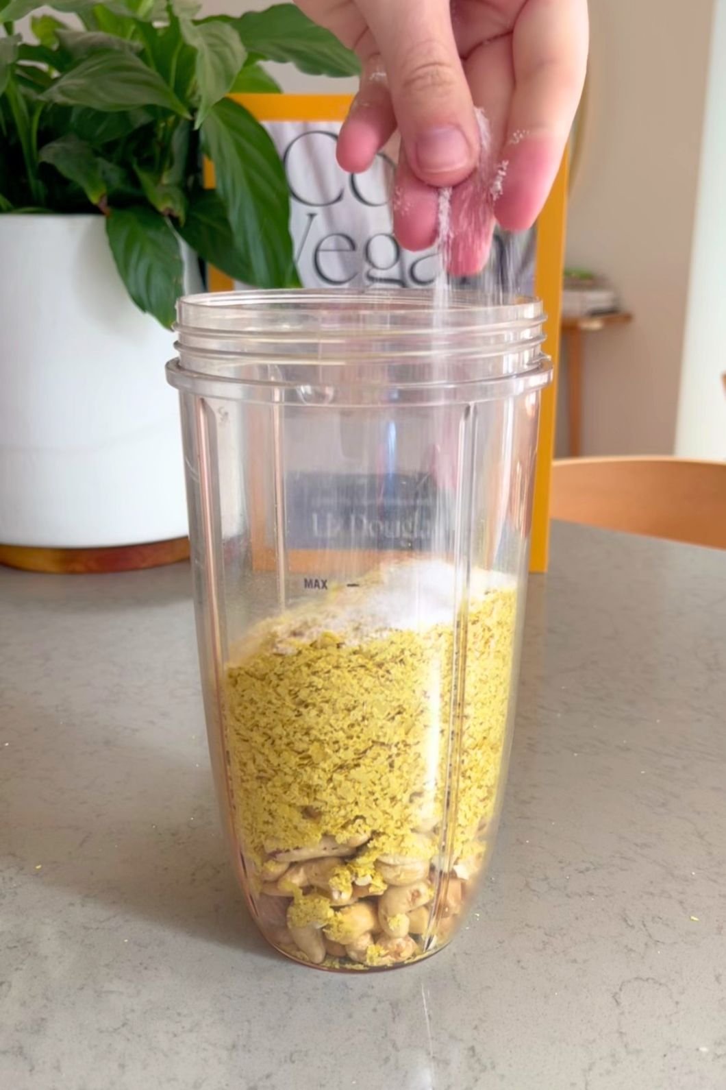 Hand sprinkling salt into a blender cup filled with crushed cashews and yellow flakes.