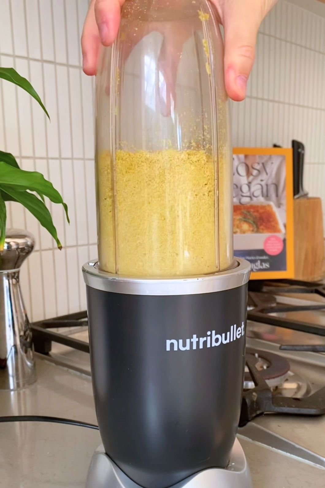 Hands holding a blender cup of coarse yellow mixture seated on a NutriBullet base.