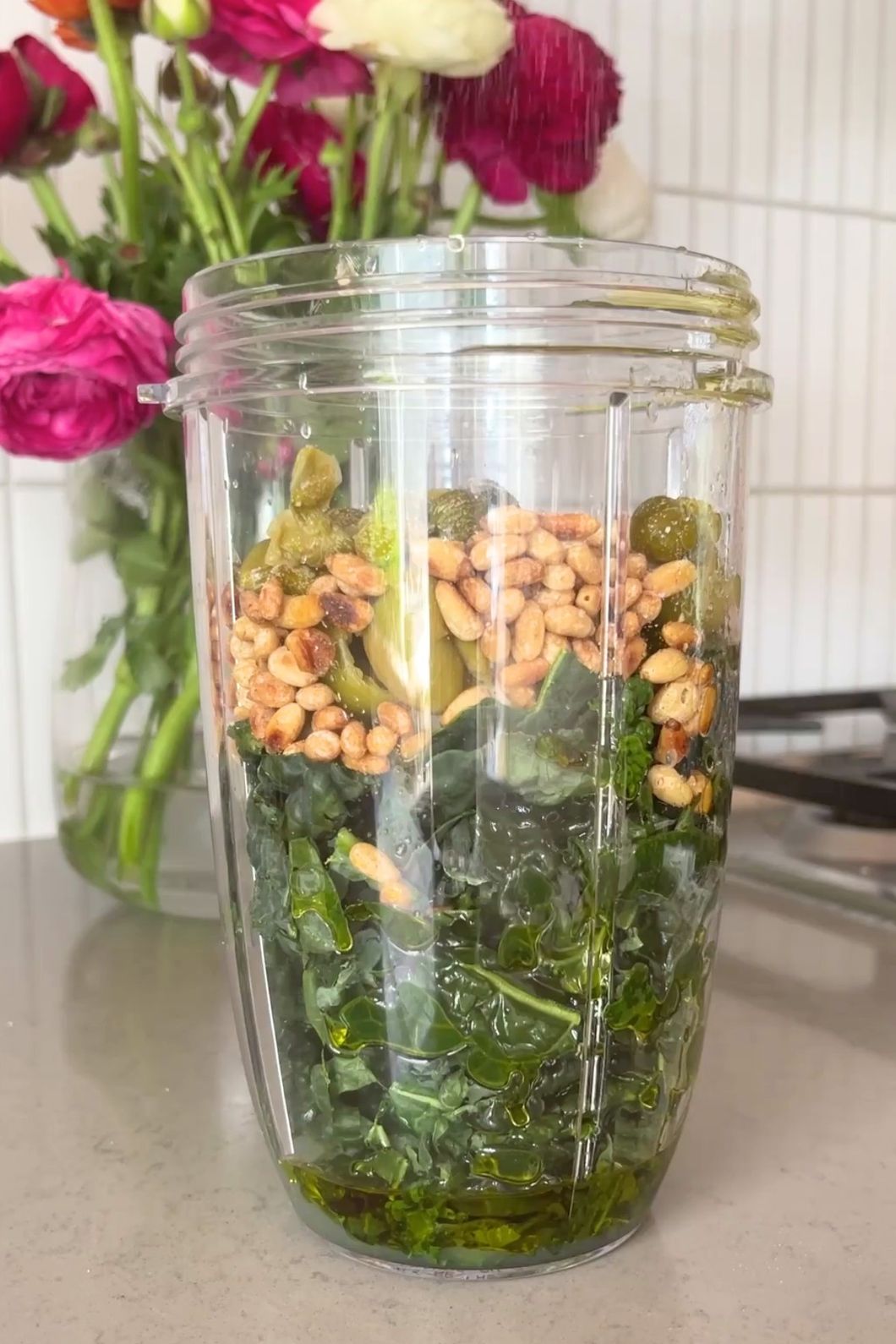 Chopped kale layered with pine nuts, green olives, and olive oil in blender cup.