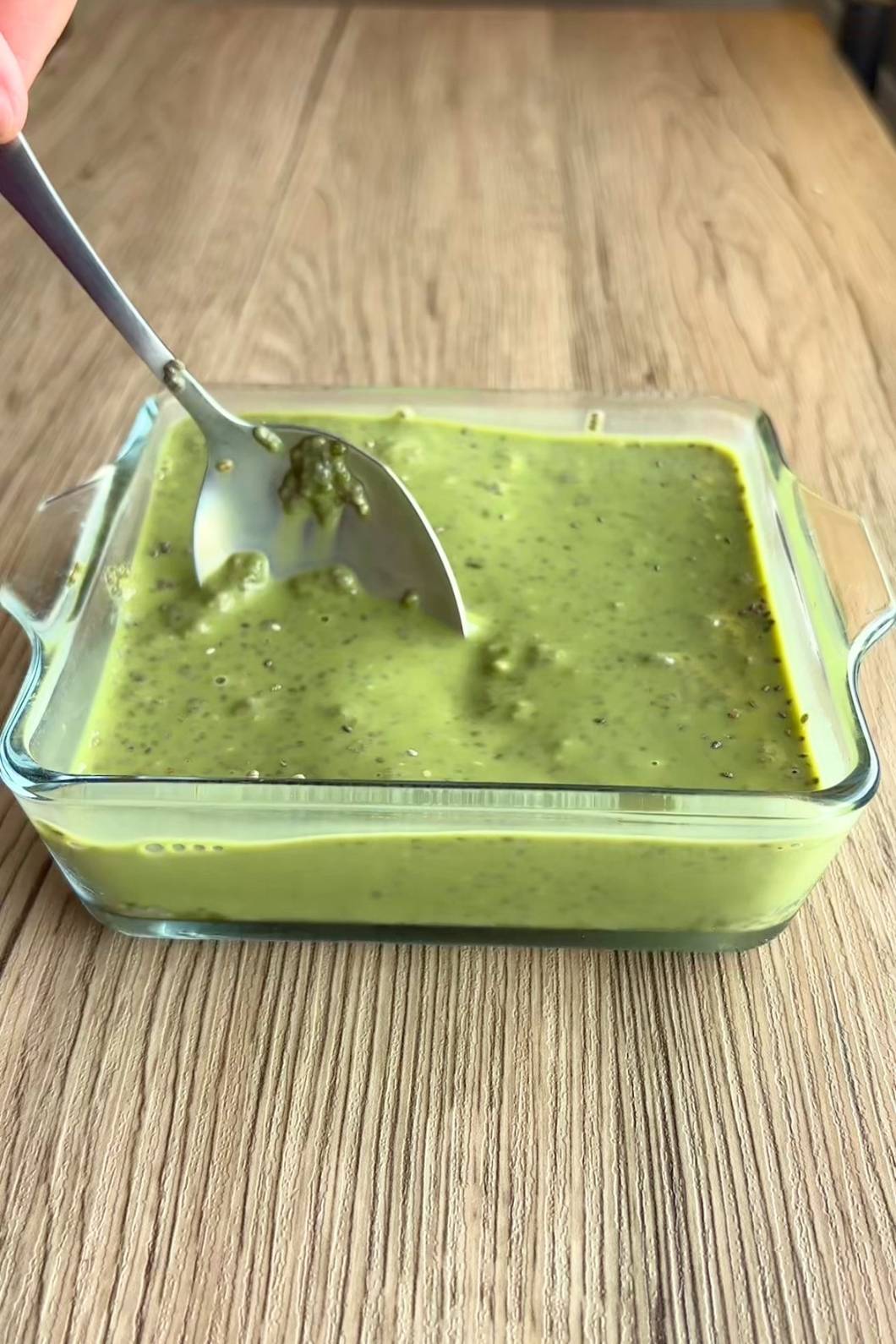 Spoon stirring thick speckled green pudding in a clear rectangular glass dish.